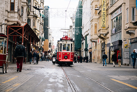 Tram traveling on road surrounded by beige commercial buildings with people walking on both sides during daytime thumbnail