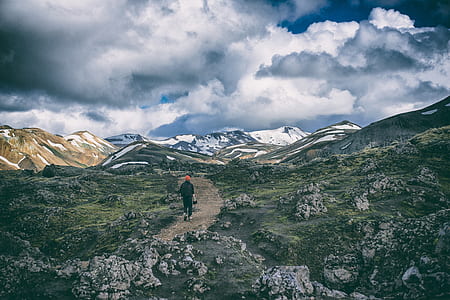 person wearing black top walking on brown road surrounded with gray rocks and green grass under white clouds and blue sky during daytime thumbnail
