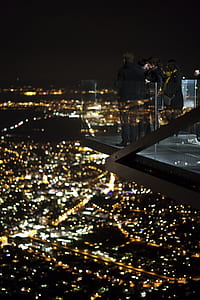 four people standing on deck with glass railings facing city with lights during nighttime photo thumbnail