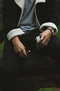 man in black jacket holding blue flip-top cigarette box and gray flip lighter lighted thumbnail