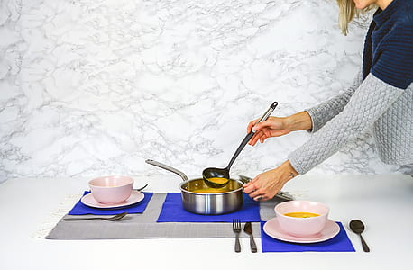 woman in blue and grey sweater pouring soup to pink bowls from a grey cooking pot on white table thumbnail