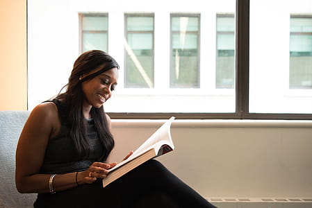 Woman Wearing Black Crew-neck Sleeveless Top Sitting of Gray Sofa While Reading Book thumbnail