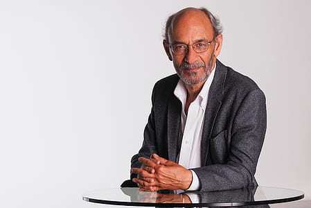 man wearing black notched lapel suit jacket leaning on clear glass-top table thumbnail