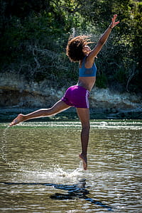 Woman in Blue Sports Bra and Purple Shorts Leaping Above Body of Water thumbnail