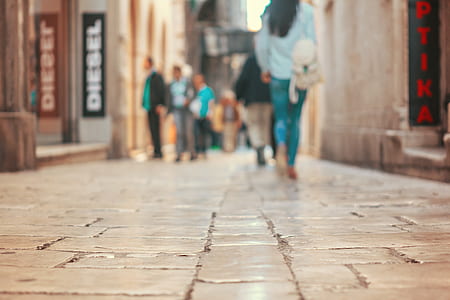 tilt lens photography of woman wearing blue jeans and blue shirt walking on brick pavement thumbnail