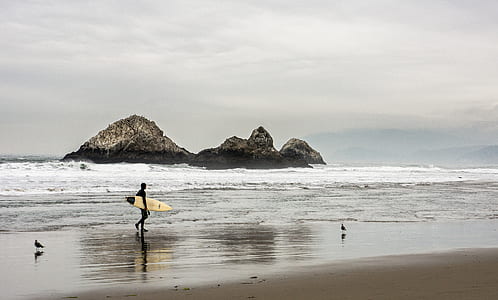 Man Carrying Surfboard in Front Bodies of Water on Brown Sand thumbnail