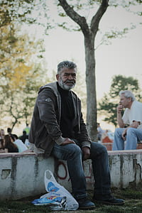 Man Wearing Gray Jacket Sitting on White Stone in Park during Day Time thumbnail