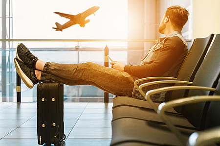man sitting on airport gang chair while feet on luggage bag thumbnail