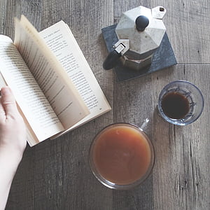 person holding book beside two clear glass cups beside gray moka pot thumbnail