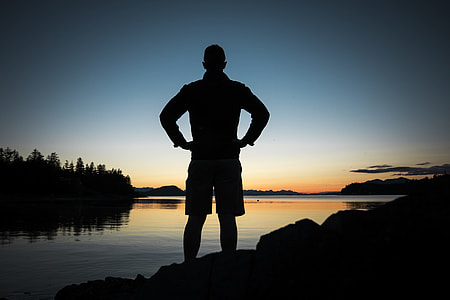 silhouette of man standing on stone near the calm body of water during sunrise thumbnail