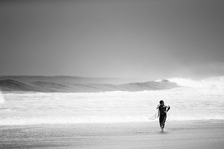 gray scale photography of man holding surfboard on seashore thumbnail