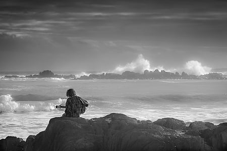 grayscale photography of man holding guitar sitting on pile of stones thumbnail