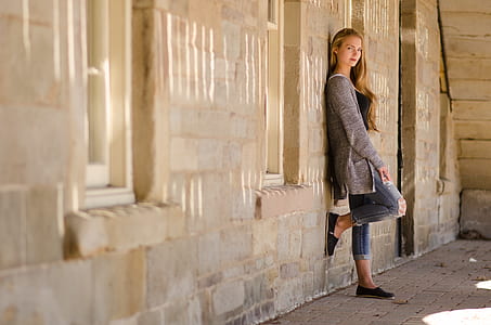woman wearing gray cardigan and blue denim jeans leaning on gray wall thumbnail