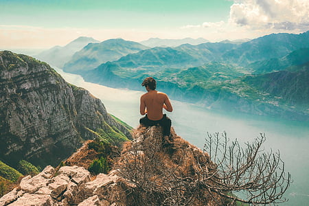 topless man sitting on rock in front of cliff overlooking hills and body of water under white cloudy skies at daytime thumbnail