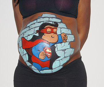pregnant woman with superman paint on tummy standing beside white wall thumbnail