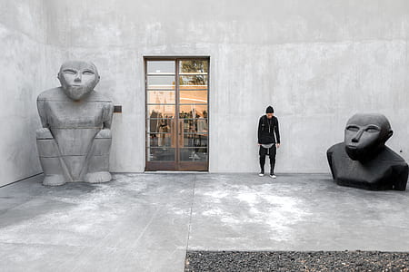 man standing beside gray wall between statues thumbnail