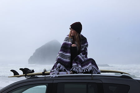 woman with blanket sitting on surfboard on top of vehicle near sea in distant of mountain isle thumbnail