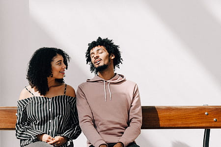 man in pullover hoodie sitting on bench while closing his eyes behind woman looking at him while smiling with white backdrop thumbnail