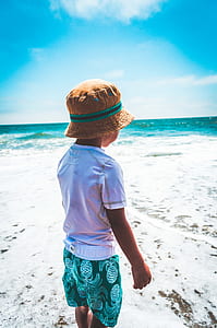 child wearing white t-shirt, green and white pineapple print shorts and khaki bucket hat over white sand seashore across wavy body of water during daytime thumbnail