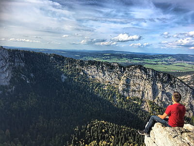 man sitting alone on rock cliff facing pine trees, mountain, and cloudy sky thumbnail
