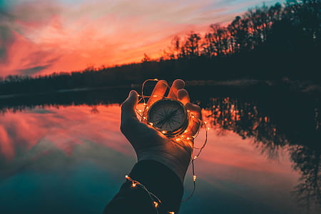 person holding compass and string light near body of water during sunset thumbnail