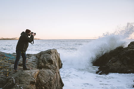 man on top of cliff taking picture of sea wave smashing on the rocks during daytime thumbnail