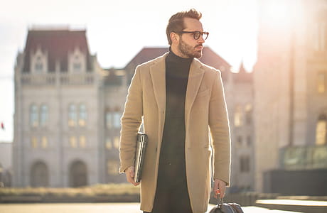 Young white bearded male wearing black turtleneck top with long overcoat and eyeglasses carrying a laptop,as he walks on the street thumbnail