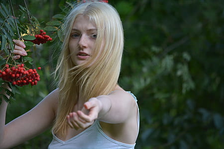 woman wearing white spaghetti strap top reaching out left hand and right hand holding bunch of red berries thumbnail
