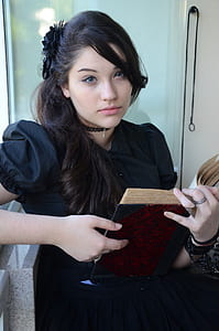 Woman With Black Hair and Wearing Black Cap Sleeve Dress Holding Book With Two Hands thumbnail