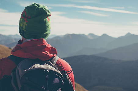 person wearing red jacket, green knit cap and backpack facing on mountain during daytime thumbnail