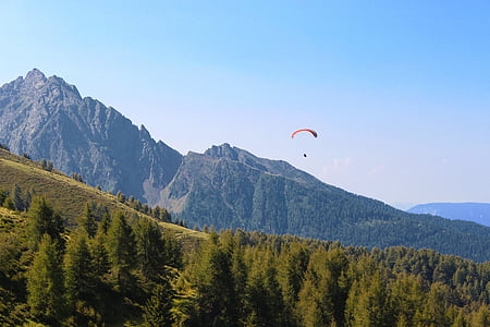 person parachuting down hillside with pine trees under blue sky during daytime thumbnail