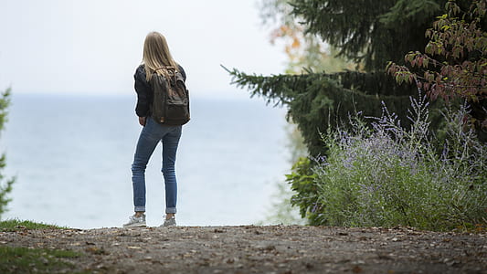 woman in black jacket and blue jeans wearing backpack standing near plants thumbnail