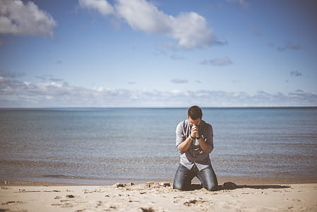 man wearing grey shirt kneeling down on beach shore praying under cloudy sky thumbnail