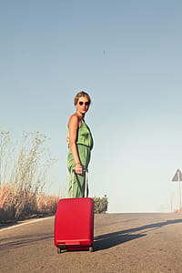 A young blonde woman searing olive green dress and sun glasses posing with red luggage Trolley Bag thumbnail