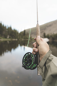 man holding brown and green fishing rod standing near lake under white sky thumbnail