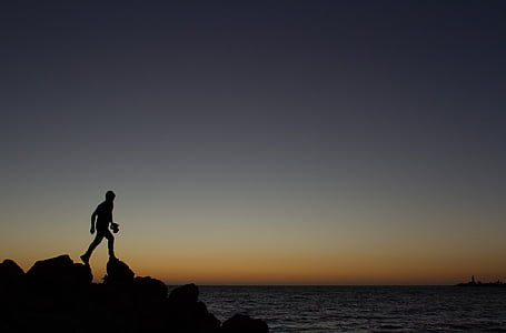 silhouette of person walking on rock at the edge of ocean during golden hour thumbnail