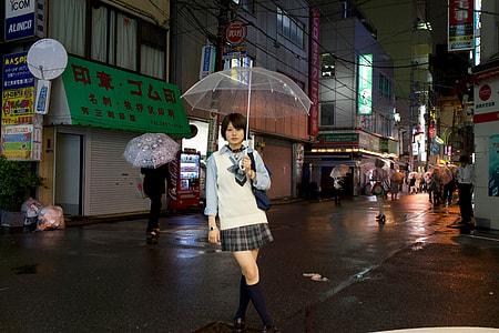woman holding clear umbrella wearing white and blue collared long-sleeved shirt with black and gray plaid mini skirt standing surrounded by buildings during night thumbnail