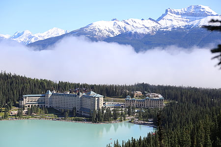 gray concrete building next to body of water and green pine trees across ice cover mountain thumbnail