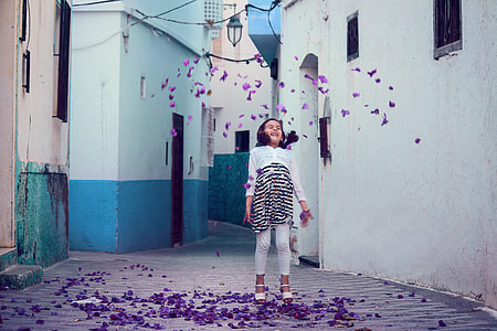 girl in white and black striped long-sleeved shirt and white pants standing in front of white painted wall with purple petals thumbnail