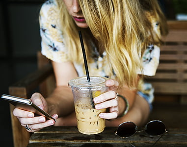 selective focus photography of blonde haired woman sipping yellow liquid while using smartphone thumbnail