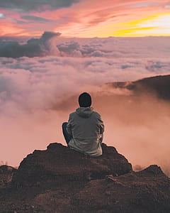 man in grey jacket looking at clouds while sitting on brown cliff thumbnail