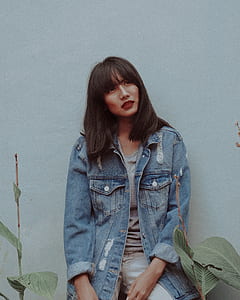 sitting woman wearing blue denim stonewashed button-up jacket, gray shirt and white bottoms leaning behind white wall in between green plants thumbnail
