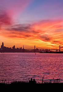 silhouette of couple sitting near body of water with buildings and bridge in the background thumbnail