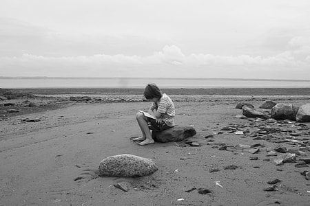 grayscale photo of girl reading book on beach while sitting on rock thumbnail