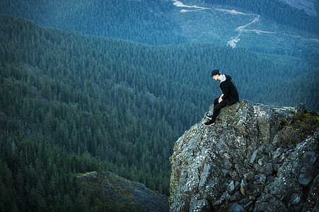 man sitting on rock formation watching green trees during daytime thumbnail