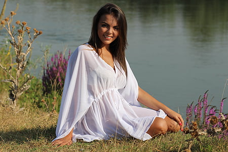 smiling woman wearing white v-neck flare-sleeved cover-ups sitting on green grass beside body of water during daytime thumbnail