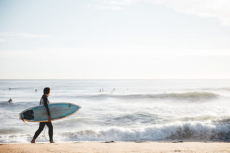 A young Caucasian surfer carrying surfboard on the beach with high waves in the background thumbnail