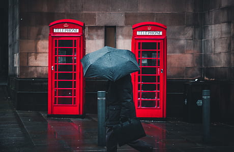 person wearing coat holding umbrella walking beside red telephone booths thumbnail