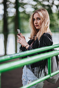 Blonde woman in a black jacket and ripped jeans by a green handrail thumbnail