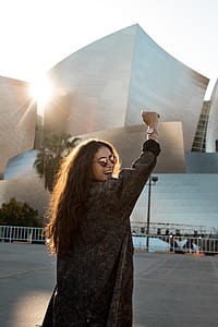 smiling woman wearing brown jacket and sunglasses in front of white concrete building thumbnail